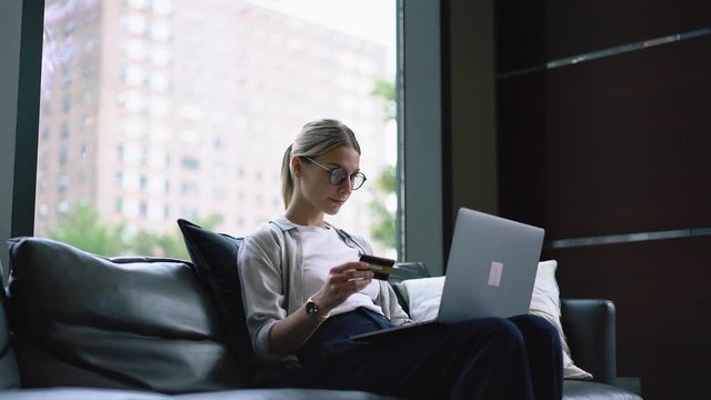Hipster blogger holding plastic card and typing number on keyboard of modern laptop using wireless internet connection for booking tickets on website at home apartment with stylish interior

