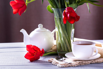 A cup of tea, a white kettle and red tulips in a vase on the wooden table.
