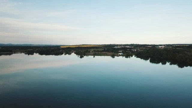 Shot of a drone on a clear water surface. Calm lake without waves and reflecting water. Water tank from a bird's eye view. A biotope that is filmed after the golden hour. Czechia, South Moravia