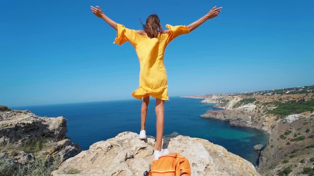 Full length epic slow motion of taned traveler woman standing on top rock cliff with open arms in front of amazing seascape, bright yellow dress and hair blowing in the wind.