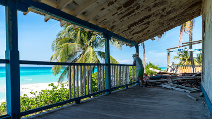 A man sitting on the patio of an old decayed house on a tropical beach.
