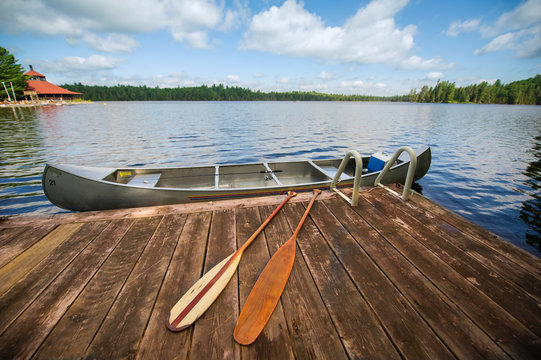 Canoe tied to a wooden dock on a summer day. On the dock there are two paddles. There's a fishing rod inside the canoe. 