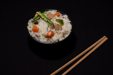 A bowl of rice with vegetables and soya chunks along with chopsticks in a black copy space background. Food photography.