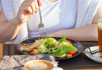 Caesar salad on a dark plate and the hands of a girl in a white T-shirt, which is going to eat a salad, with a fork. A plate on the table and two coffees.