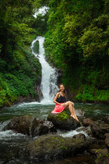 Happy smiling Caucasian woman sitting on the rock and enjoiyng waterfall landscape. Travel concept. Dedari waterfall in Sambangan, Bali.