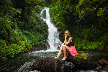 Young Caucasian woman sitting on the rock and enjoiyng waterfall landscape. Travel concept. Dedari waterfall in Sambangan, Bali.