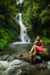 Fototapeta na wymiar Attractive Caucasian woman sitting on the rock and enjoiyng waterfall landscape. Travel lifestyle. Dedari waterfall in Sambangan, Bali.