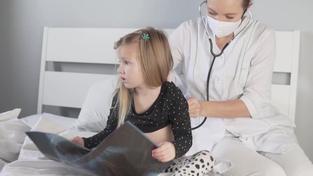 The doctor listens to a small patient with a stethoscope and shows her a picture of her lungs during the coronavirus epidemic.