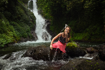 Fototapeta na wymiar Young Caucasian woman sitting on the rock and playing with water in the river near the waterfall. Travel lifestyle. Dedari waterfall in Sambangan, Bali.
