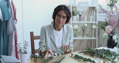 Young business woman fashion decor designer or florist making flower arrangement sitting at desk in studio. Creative occupation specialist doing handmade stylish eucalyptus floral composition design.