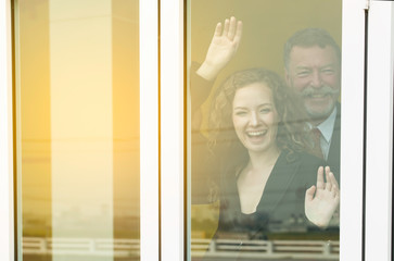 Fototapeta na wymiar Business middle aged man and beautiful woman behind their glass window of home say hello to others people,Happy old man and young woman in suit standing and gesturing raised up hands.Work form home.