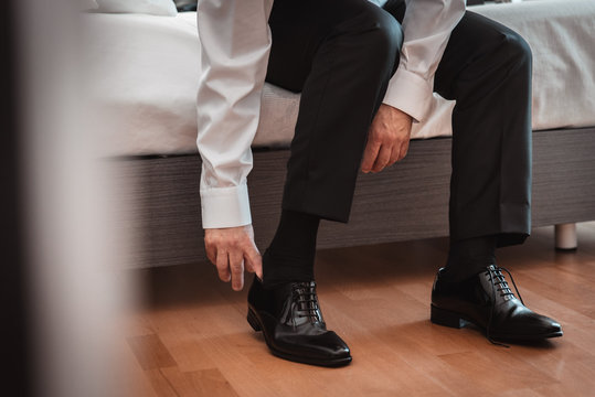 groom in white shirt and black pants sitting on the bed putting on black shoes for the wedding