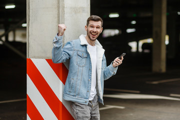 European young man in a fashion jacket in a denim jacket holds a phone posing near a pillar with a red-white line in a city parking lot.