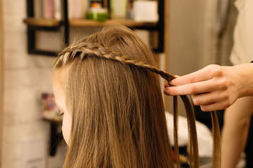 Naklejka premium Process of braiding the master weaves braids on her head blond little girl in beauty salon close up. Professional hair care and creating hairstyles.