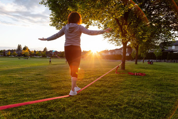 Girl walking on a Slackline in a park during a sunny sunset. Taken in Fraser Heights, Surrey, Greater Vancouver, British Columbia, Canada.