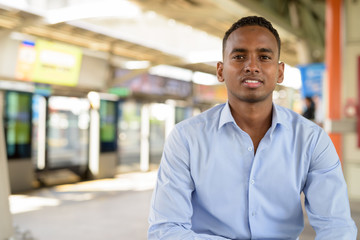 Happy young handsome African businessman smiling while sitting at the sky train station - obrazy, fototapety, plakaty