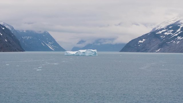 Iceberg floating between two snow capped mountains in Prince Christian sound in Greenland at 120fps slow-motion filmed from the front of a cruise ship with moody dark clouds in the distance