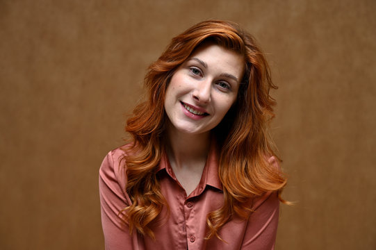 Photo taken in the studio on a beige background. Close-up portrait of an actress Caucasian woman with long red hair showing happy emotions. The model is wearing a red shirt.