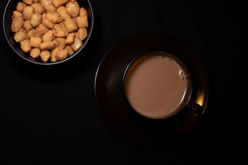 Top down image of infused tea in a transparent glass cup and soccer along with a bowl of snacks kept in black copy space background. Indian beverages and food photography.