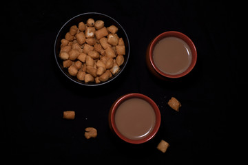 Top down image of infused tea in earthen cups along with a bowl of snacks kept in black copy space background. Indian beverages and food photography.