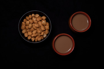 Top down image of infused tea in earthen cups along with a bowl of snacks kept in black copy space background. Indian beverages and food photography.