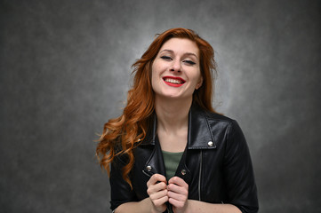 Pretty model actress posing with different emotions on a gray background in the studio. Studio photo of a young caucasian woman with long red hair in a black jacket. - 348422380