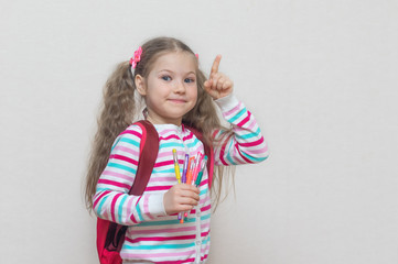 Back to school. Portrait of a blonde schoolgirl in a jacket with colored gel pens in her hands. The girl is smiling and looking at the camera. Thumbs up. Light background. Education. copy place.