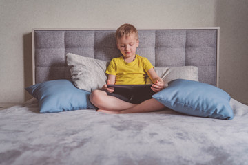 Distance education, learning. Boy is sitting on the bed with a tablet in the room. Child with a gadget. Teenage child using tablet pc.