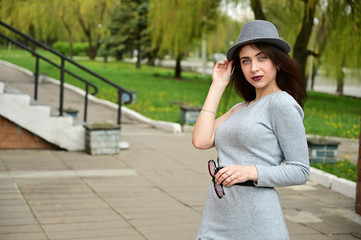 Photo of a cute young caucasian brunette girl in a hat in a gray dress with glasses in her hands. Model posing in a city park outdoors