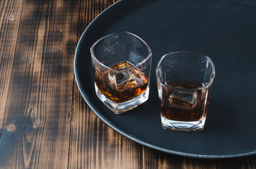 Glasses of whiskey with ice cubes on a black tray and wooden table/Two Glasses of whiskey with ice cubes on a black tray and wooden table with copyspace.