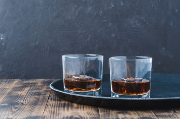 Glasses of whiskey with ice cubes on a black tray and wooden table/Two Glasses of whiskey with ice cubes on a black tray and wooden table. Black background with copyspace.