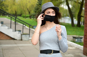 Photo of a cute young caucasian brunette girl in a hat in a gray dress wearing a black medical mask. Model posing in a city park outdoors