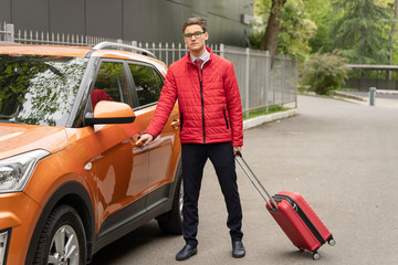 Portrait of enterpriser in a red jacket traveling with modern strolley case by car. Beautiful stylish businessman travel with luggage Transport and transfer concept.