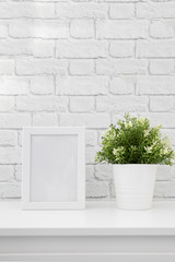 White frame mockup with green plant on a white table against white brick wall. Empty poster frame mockup for design.