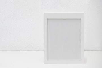 White frame mockup  on a white table against white wall. Empty poster frame mockup for design.
