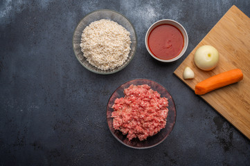 Ingredients for meatballs - minced meat, rice, tomato paste, onion, garlic, carrots on a cutting board on a dark background. Top view