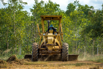 Motor grader clearing and leveling construction site surface with forest in the background. Grader industrial machine on road construction work.