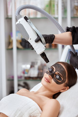 pretty girl with carbon mask on face receiving laser procedures in black glasses by cosmetologist in spa salon