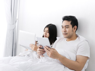 Happy Asian couple using tablet and laptop on bed, work from home, stay home stay safe concept.