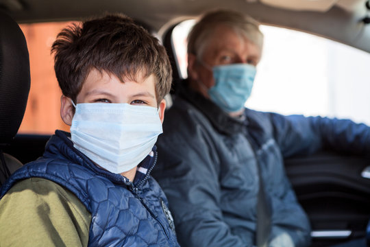Preteen age boy sitting on passenger seat with his father as driver, men wearing surgical masks against coronavirus, looking at camera