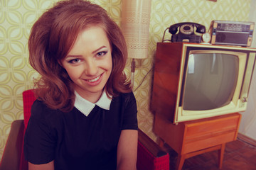 Young smiling ecstatic woman looking at camera and happy smiling in room with vintage wallpaper and interior, retro stylization 60-70s. Furniture, tv set and another technique of the mid 20th century