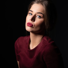 Charming young woman with blue eyes and long fair hair posing at studio over dark background. Amazing stylish girl with perfect makeup wearing elegant deep red dress. Beauty and fashion