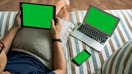Man's hands is holding tablet with green screen. There are a laptop and smart phone with green screen on sofa, work from home, online meeting, e commerce concept.