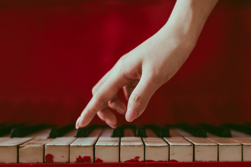 a close-up of a side view with female hand with pink manicure playing the piano. An old red piano with black and white keys. A palm above the keyboard. Graceful fingers of composer or musician
