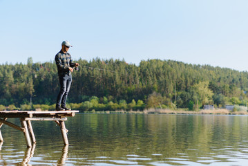 Fototapeta na wymiar Fishing for pike, perch, carp. Fisherman with rod, spinning reel on river bank. Man catching fish, pulling rod while fishing on lake, pond with text space. Wild nature. The concept of rural getaway.