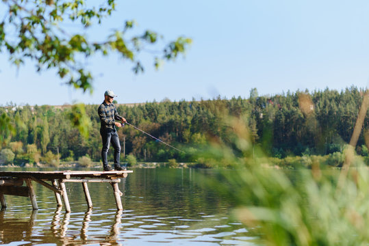 Fishing for pike, perch, carp. Fisherman with rod, spinning reel on river bank. Man catching fish, pulling rod while fishing on lake, pond with text space. Wild nature. The concept of rural getaway.
