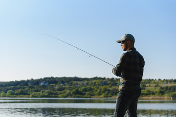 Fishing for pike, perch, carp. Fisherman with rod, spinning reel on river bank. Man catching fish, pulling rod while fishing on lake, pond with text space. Wild nature. The concept of rural getaway.