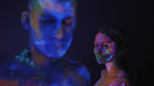 Portrait of a couple with UV drawings on the skin in the dark under the light of fluorescent lamps, the man in the foreground blurred. Body art on the couple's body glows in neon light.
