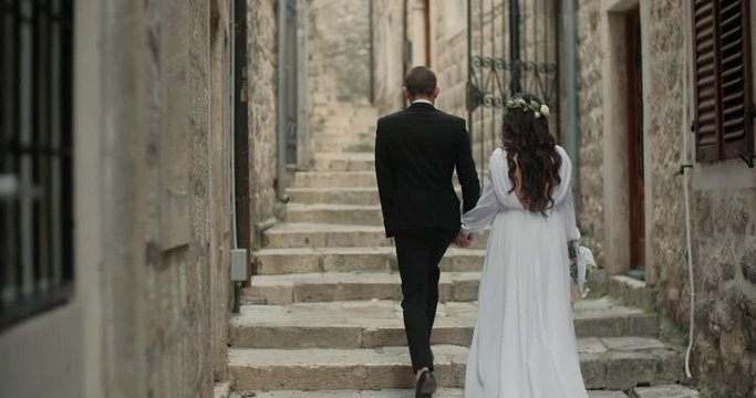 Back view of bride in beautiful white dress and groom walking in Europe old town, romantic wedding couple holding hands together in Montenegro.
