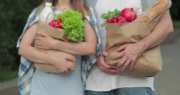 Crop view of girl and guy holding paper craft bags full of food and walking at street. Couple in casual clothes carrying their shopping in hands. Concept of healthy lifestyle